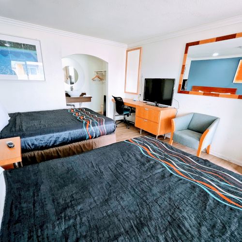 A hotel room with two double beds, a wall-mounted TV, a desk, a chair, and a large mirror. The room is neatly arranged and bright.