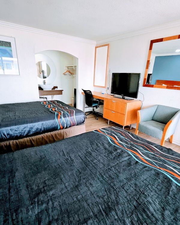 A hotel room with two double beds, a wall-mounted TV, a desk, a chair, and a large mirror. The room is neatly arranged and bright.