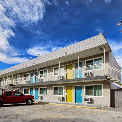 An image of a two-story motel with a red pickup truck parked in front, showing colorful doors and an outdoor staircase under a partly cloudy sky.