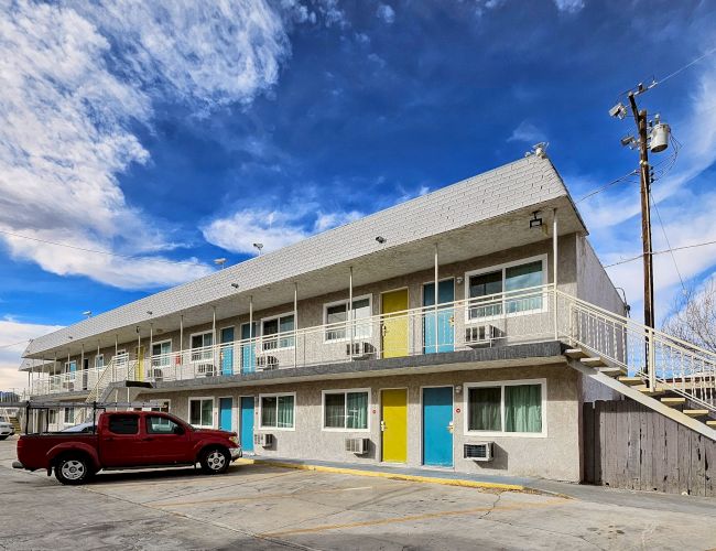 An image of a two-story motel with a red pickup truck parked in front, showing colorful doors and an outdoor staircase under a partly cloudy sky.