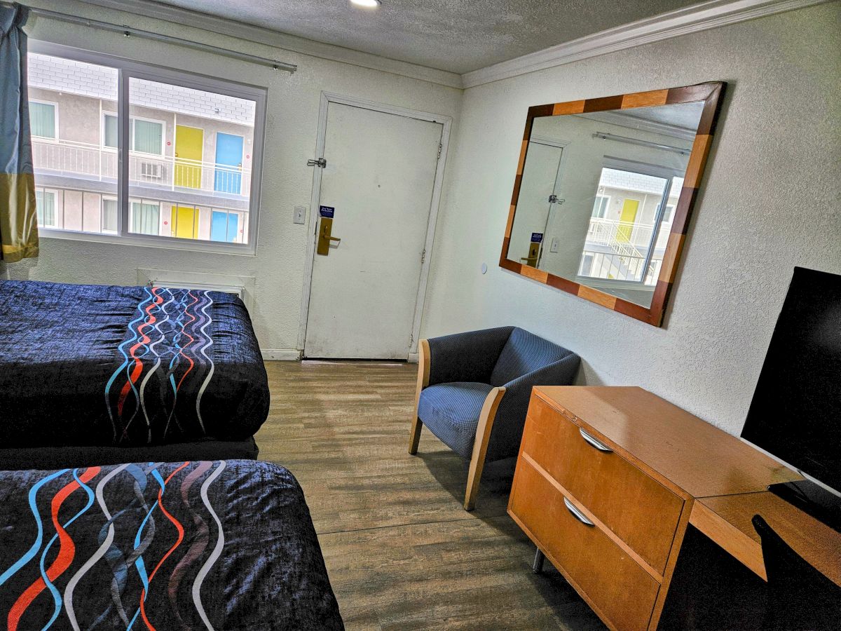 The image shows a hotel room with two beds, a large mirror, a chair, a dresser, and a TV, with a window and door visible.