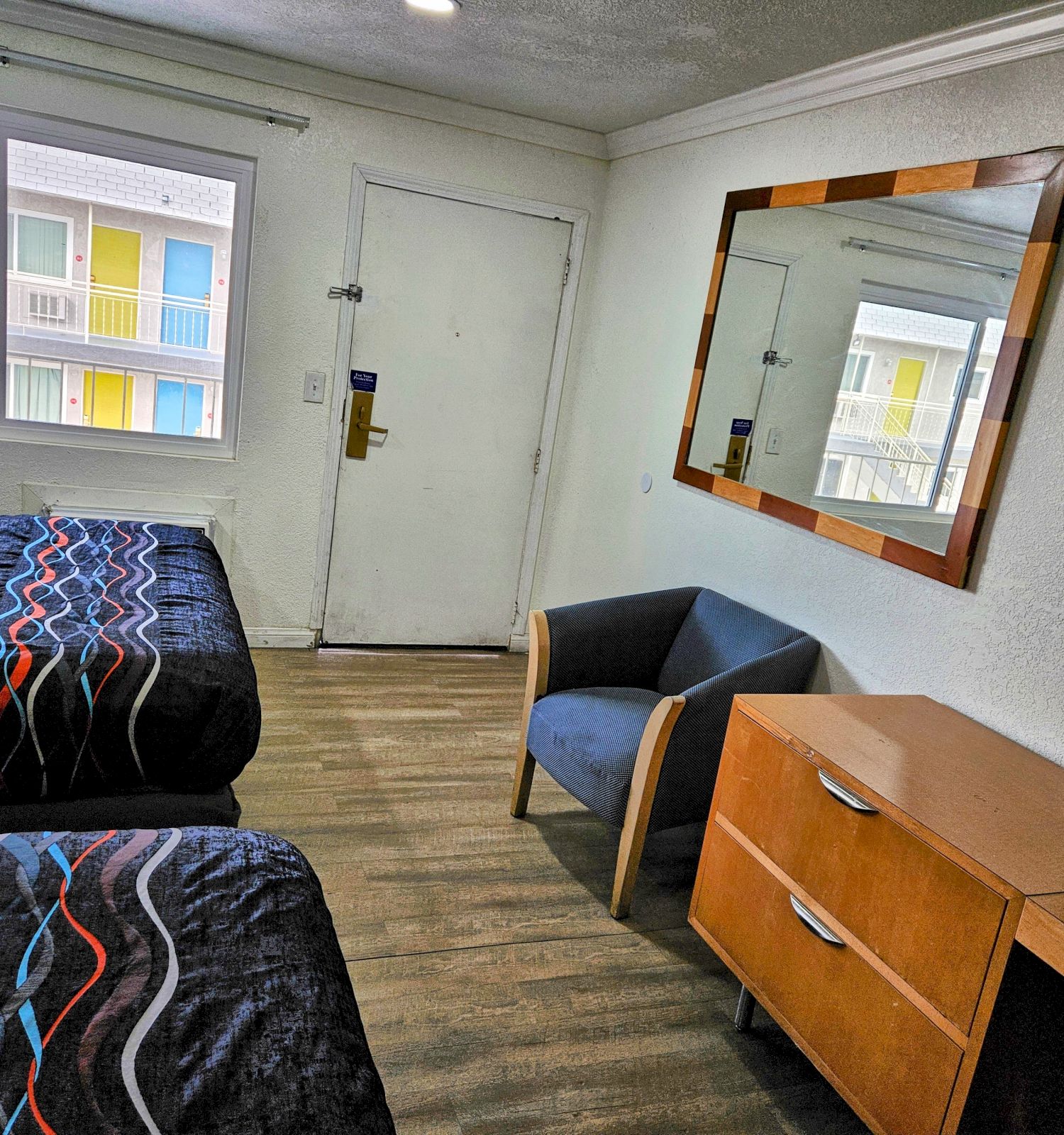 The image shows a hotel room with two beds, a large mirror, a chair, a dresser, and a TV, with a window and door visible.