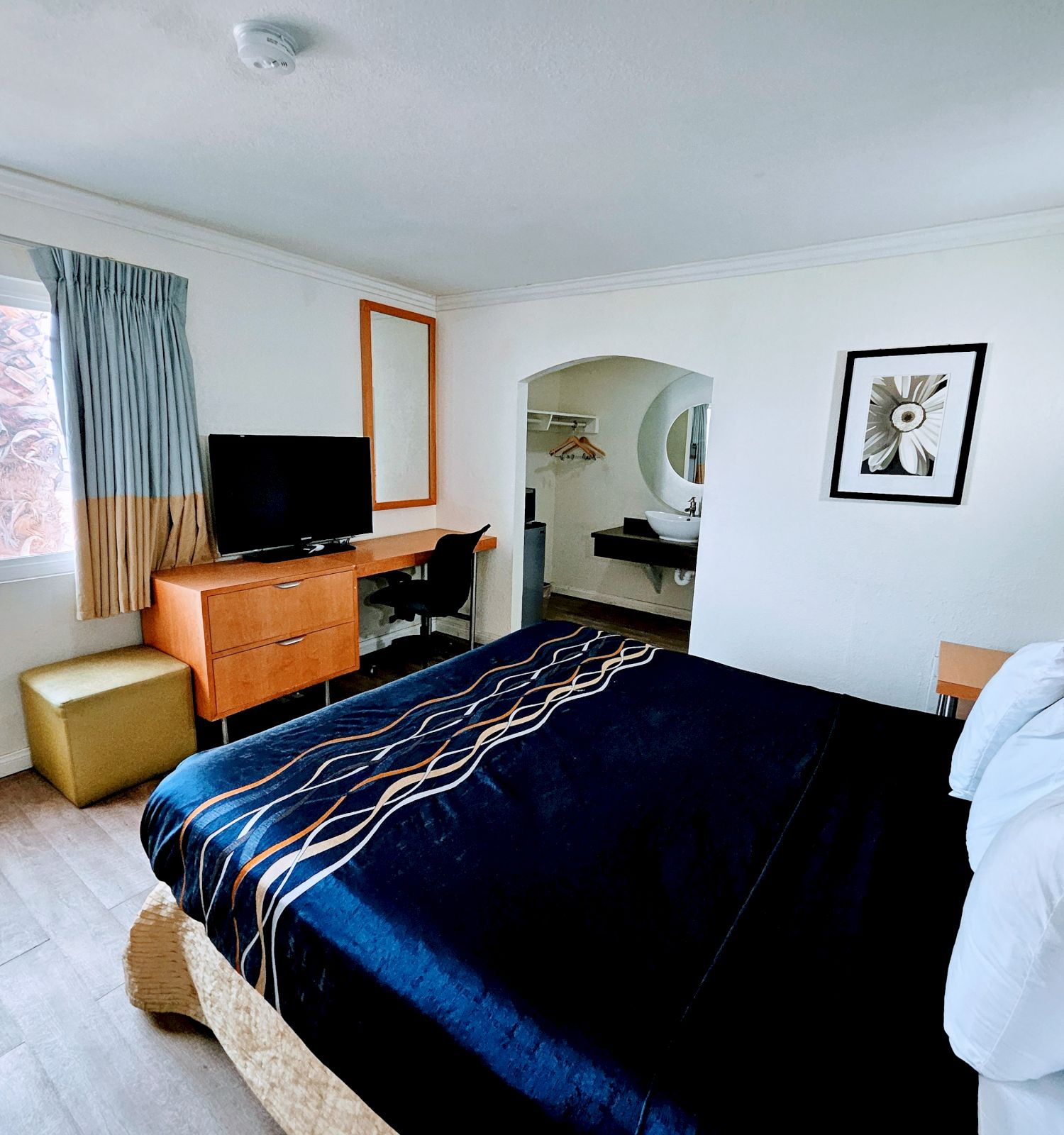 A cozy hotel room features a bed with black linens, a flat-screen TV, a wooden dresser, a window with curtains, and simple decor on the wall.