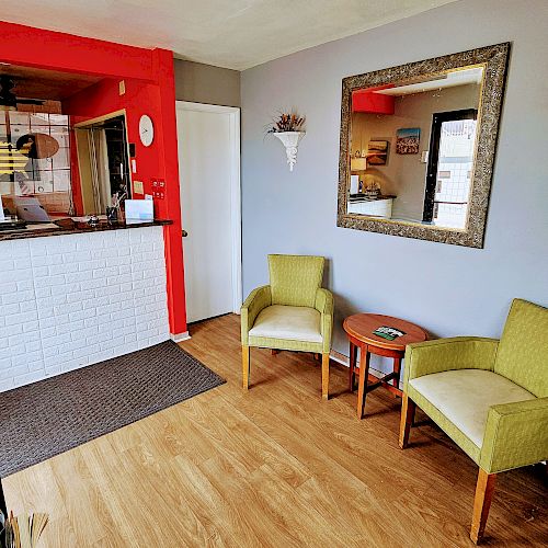 This image shows a small waiting area with a front desk, two green chairs, a small table, a mirror, and a book rack in a room with wood flooring and red walls.