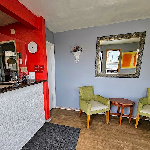 The image shows a small waiting area with two green chairs, a round table, a large mirror, a window, and a reception counter with a red accent wall.