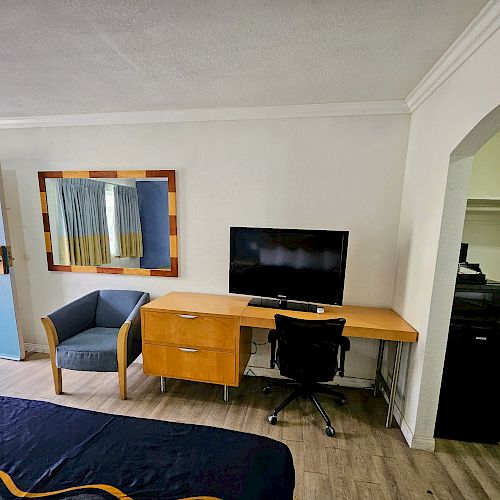 A hotel room with a bed, desk, chair, TV, and mirror; visible open door and closet area with a safe and refrigerator.