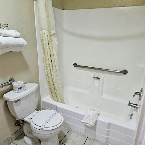 The image shows a small bathroom with a toilet, a bathtub with a shower curtain and grab bar, and a towel rack holding folded towels.