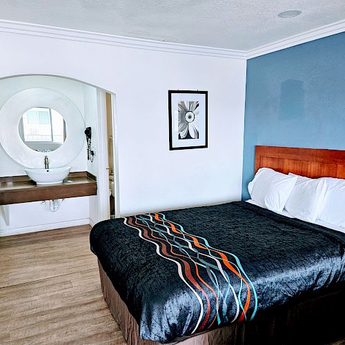 A hotel room with a bed, blue and white walls, a wall-mounted sink, artwork, and a chair is shown.