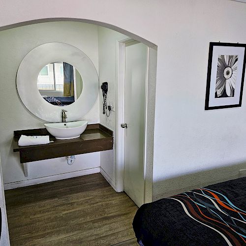 The image shows a small room with a modern sink, circular mirror, a black and white floral wall art, and a bed with a colorful bedspread.