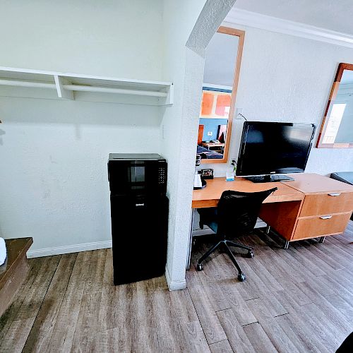 The image shows a hotel room with a desk, chair, TV, wooden floor, and a small fridge. There are hangers on a closet rack to the left.