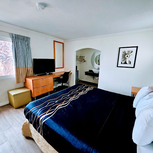 A hotel room with a king-sized bed, TV, dresser, window, artwork, and a small adjoining space seen through an archway.