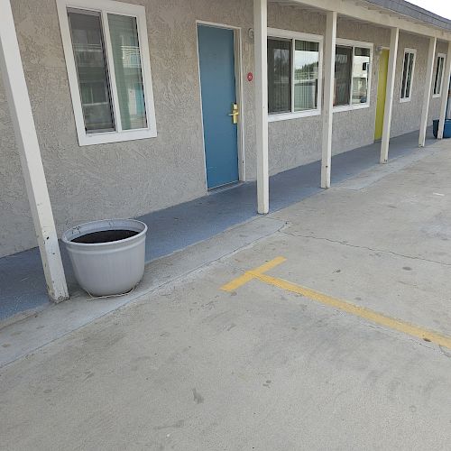 The image shows the exterior of a row of rooms, each with windows and doors, in a motel or apartment complex. A large empty planter is visible.