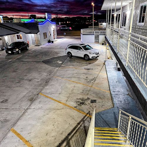 The image shows a parking area of a motel or apartment complex at sunset, with two parked cars and a staircase leading to the second floor ending the sentence.