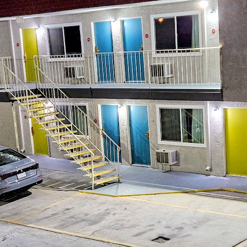 This image shows a two-story motel with a staircase, parked cars, and rooms with brightly colored doors in green and blue.