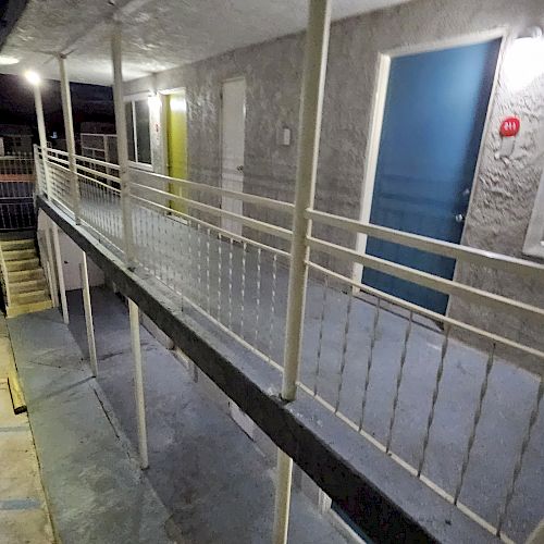 The image shows an outdoor corridor of a building with railings, illuminated by lights. There are doors in yellow and blue and a staircase leading down.