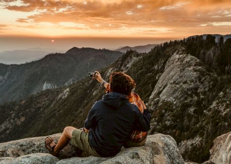 Two people are sitting on rocky terrain, taking a selfie, while overlooking a scenic mountain landscape at sunset, ending the sentence.