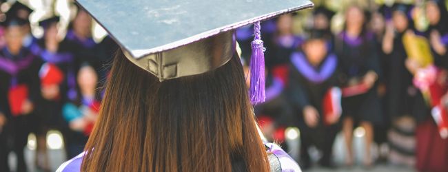 A graduate with a cap and gown stands facing a group of similarly dressed individuals, likely at a graduation ceremony.