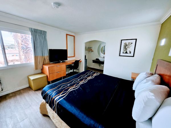 A hotel room with a bed, TV, desk, artwork, and window. There is also a small green ottoman near the window.
