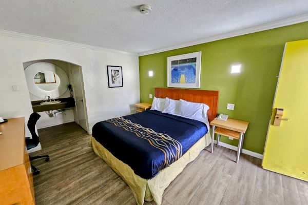 A hotel room with a bed, desk, and artwork on the wall. There is a sink area to the left with an arched opening.