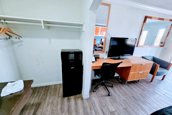 The image shows a hotel room with a closet area, mini-fridge, desk with a chair, TV, and large mirror. A window and part of a couch are visible.