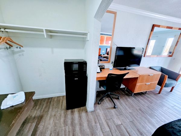 The image shows a hotel room with a closet area, mini-fridge, desk with a chair, TV, and large mirror. A window and part of a couch are visible.