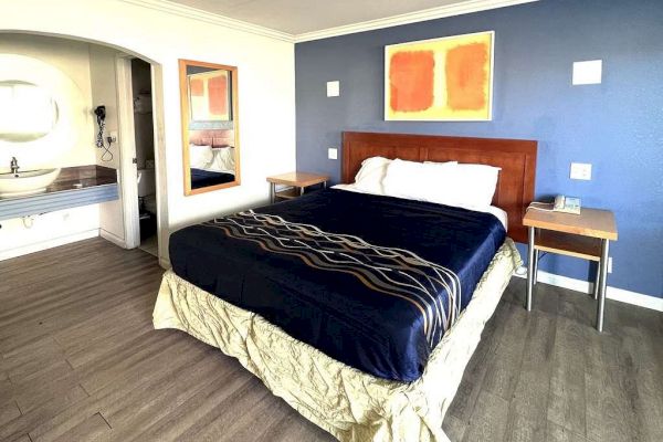 A neatly made bed with a blue and tan comforter in a modern bedroom featuring a blue accent wall, side tables, and a nearby sink area.