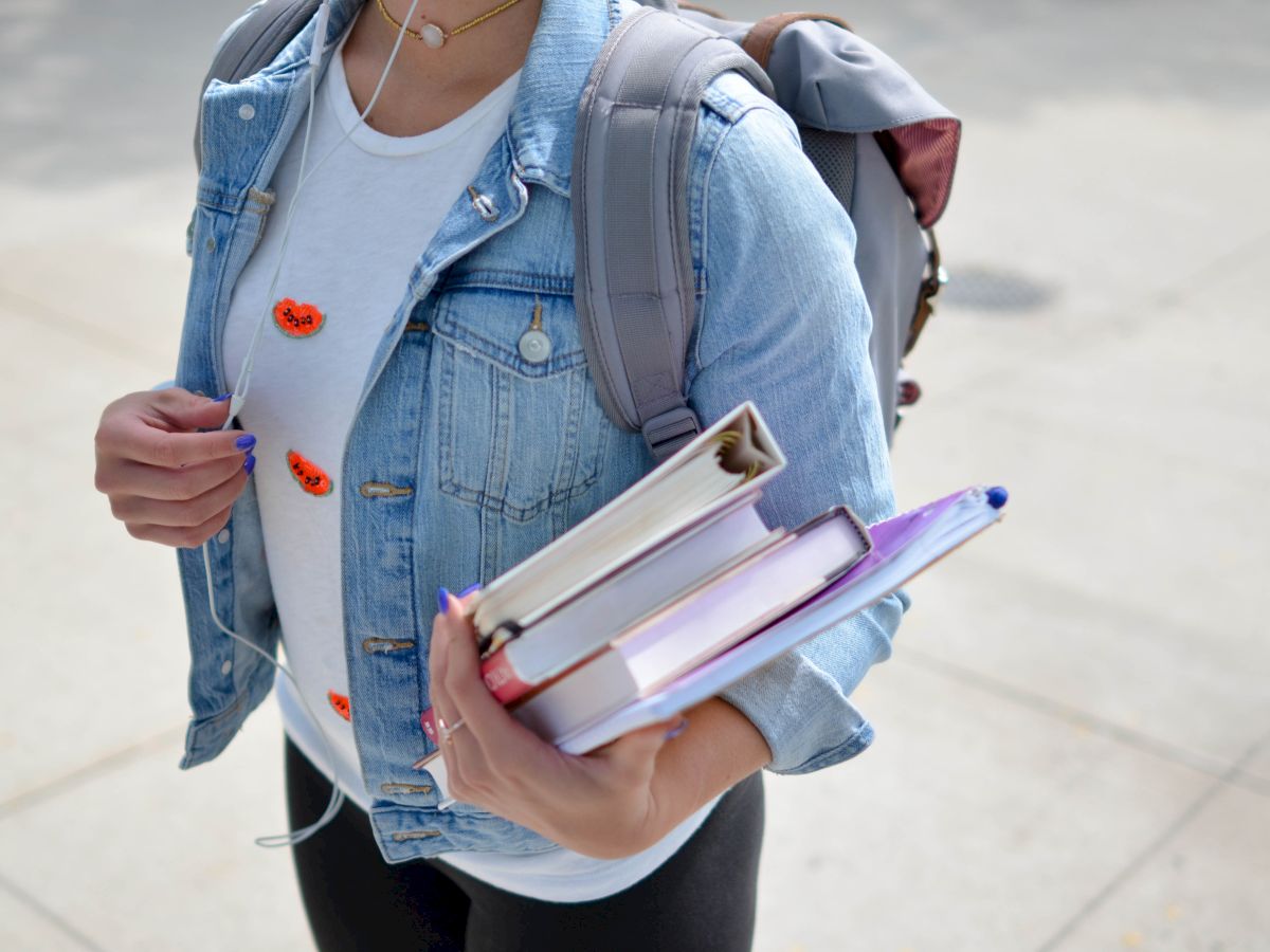 A person is holding books and wearing a backpack and a denim jacket, with earphones and a t-shirt featuring small designs on the front, standing outdoors.