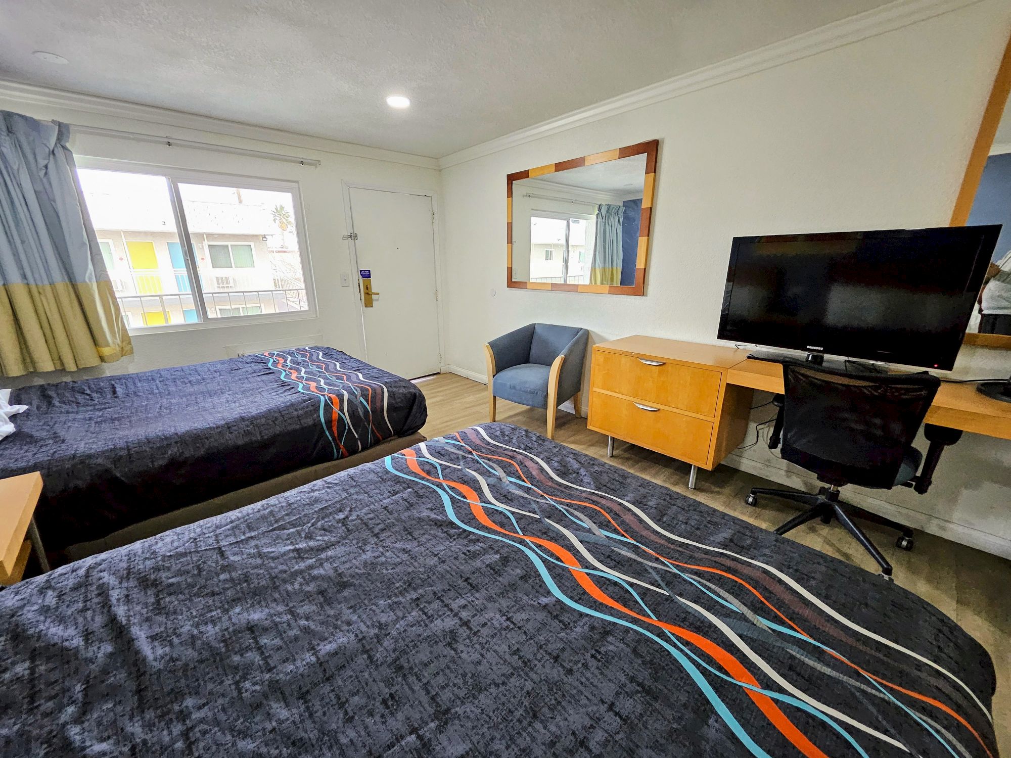 A hotel room with two beds, a TV on a desk, a chair, a large mirror, and a window with curtains, showing a building outside.