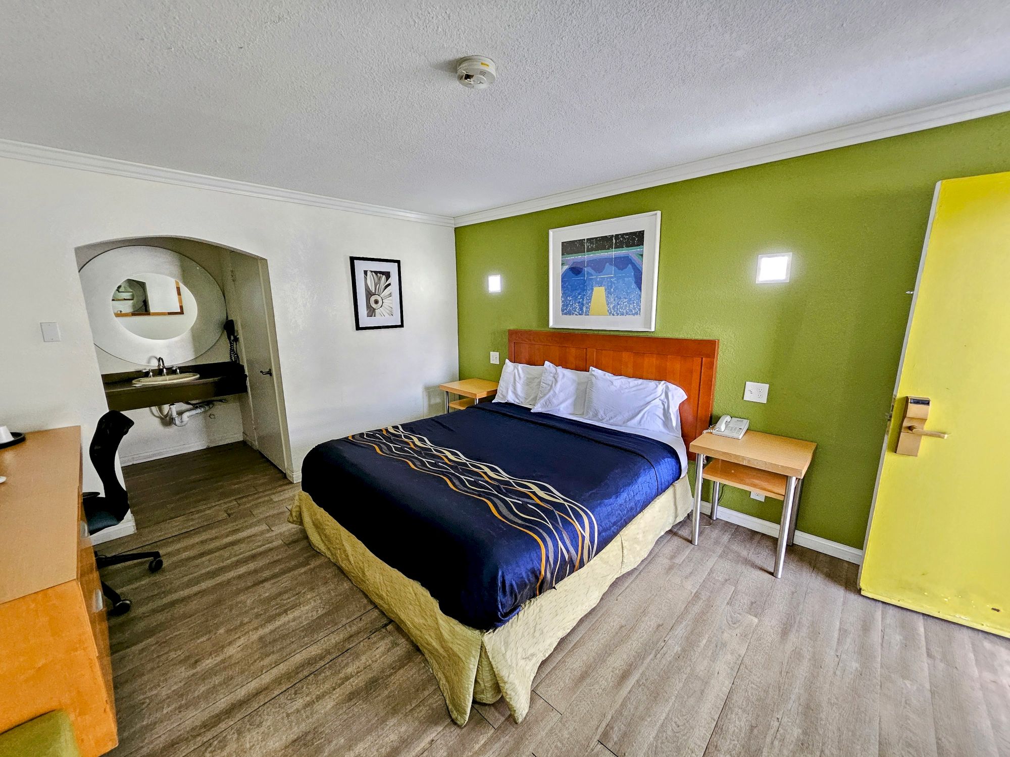 A hotel room with a double bed, a wood desk, a wall-mounted art piece, and a bathroom area in the back. A green wall and an open yellow door.